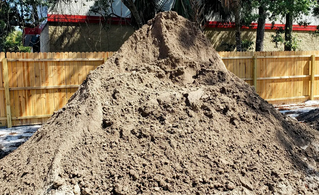 A delivered pile of sandy fill dirt beside a wood privacy fence