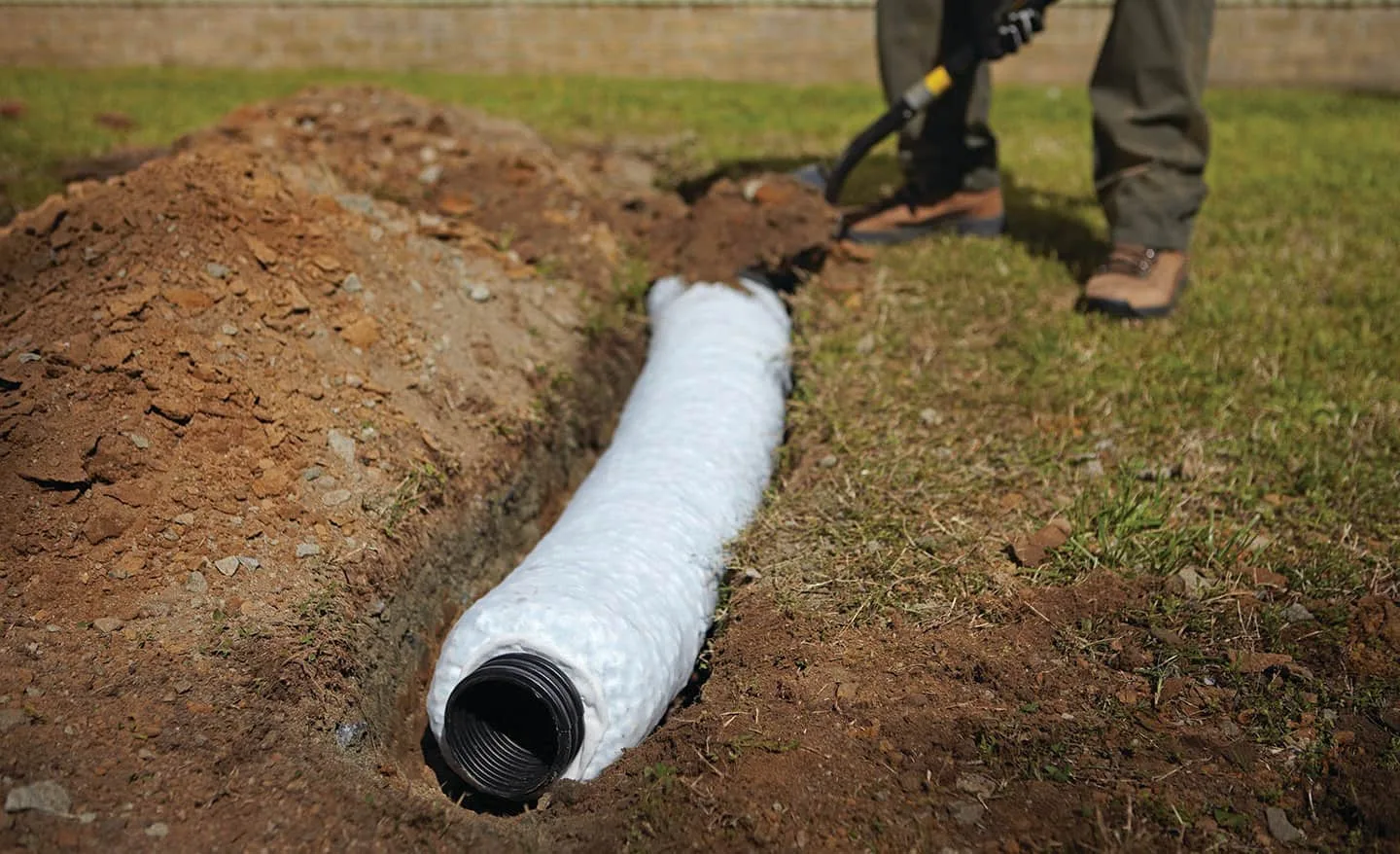 Perforated drain pipe wrapped in filter fabric being laid in an open trench during a french drain installation